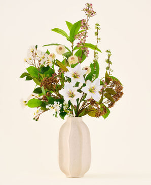 Kunstblumenstrauß aus Seide Timeless Bloom, bestehend aus einem Strauß verschiedener weißer Lilien, kleinen weißen Blumen und Grünpflanzen in einer beigen Vase mit Struktur.