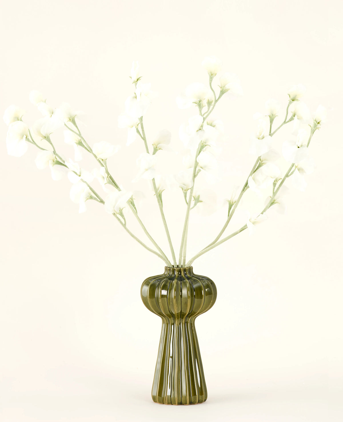 In einer grünen, gerippten Glasvase befindet sich der künstliche Seidenblumenstrauß Erbsencreme mit zarten weißen Seidenblumen und schlanken Stielen vor einem schlichten, cremefarbenen Hintergrund.