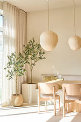 Ein heller, minimalistischer Essbereich ist mit zwei hellen Holzstühlen, einem kleinen Tisch, Papierlaternen und dem 180 cm großen künstlichen Olivenbaum ausgestattet, der einen Hauch von mediterranem Stil verleiht. Eingetopfte Zimmerbäume, durchscheinende beige Vorhänge und Sonnenlicht, das durch die großen Fenster fällt, runden diesen entspannenden Raum ab.
