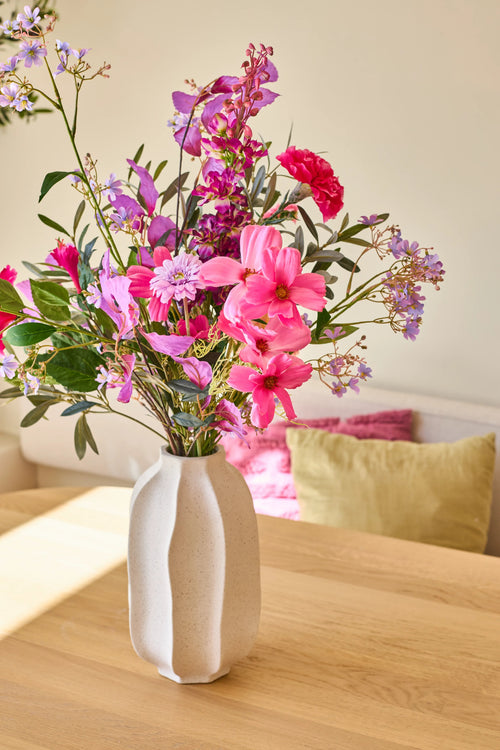 In einer weißen Keramikvase befindet sich der Strauß „Purple Dawn“ aus künstlichen Seidenblumen, ein lebhaftes Arrangement aus rosa, violetten und magentafarbenen Blumen; Die Vase steht auf einem hellen Holztisch mit bunten Kissen im Hintergrund.