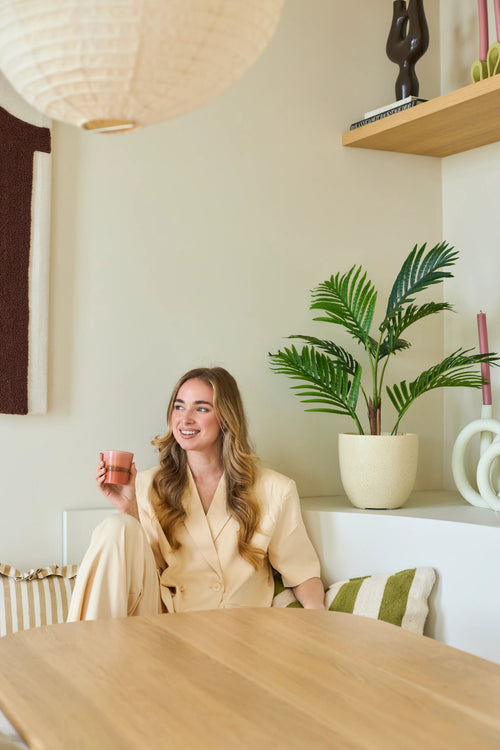Eine lächelnde Frau mit langen blonden Haaren sitzt in einem modernen, hellen Raum mit einer rosa Tasse in der Hand. Um sie herum befinden sich Grünpflanzen, darunter die üppige 85 cm große künstliche Phönixpflanze, sowie Holzregale und Dekorationsgegenstände.