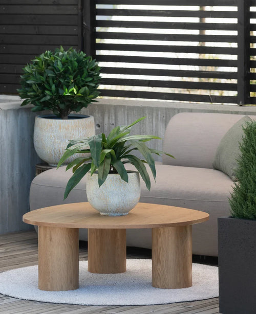Auf einem kleinen Teppich neben einem grauen Sofa auf einer Terrasse steht ein hölzerner Couchtisch mit einer 60 cm großen Kunstpflanze Bird's Deluxe darauf, im Hintergrund ein paar Topfpflanzen.