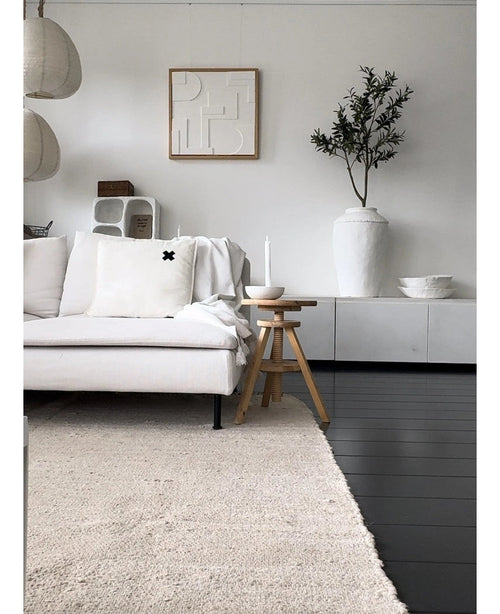 Ein minimalistisches Wohnzimmer mit einem hellen Sofa, einem neutralen Teppich, einem weißen Sideboard, einem künstlichen Olivenbaum 120 cm als Dekoelement, abstrakter Wandkunst und Holzhockern – alles in einer sanften mediterranen Atmosphäre mit neutralen Tönen.