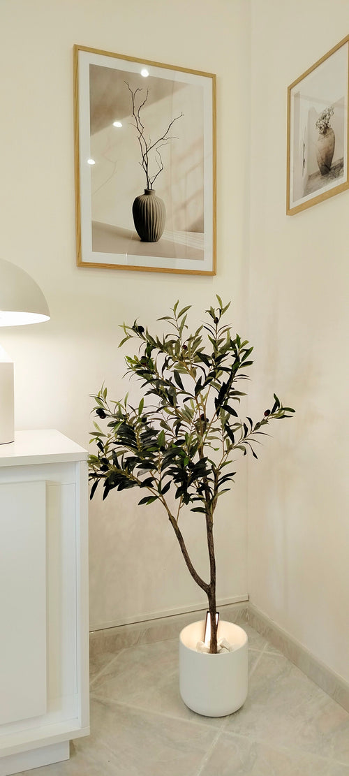 Ein künstlicher Olivenbaum 120 cm mit grünen Blättern steht in der Ecke eines hellen Raumes und sorgt für einen mediterranen Touch. Darüber hängen zwei gerahmte Kunstdrucke von Vasen mit Zweigen, während links teilweise ein weißer Schrank und eine Lampe zu sehen sind.