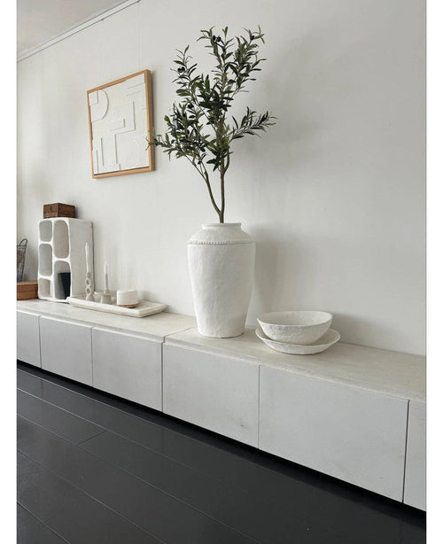 Ein minimalistischer Raum mit einem weißen Sideboard, dem künstlichen Olivenbaum 120 cm als Dekoelement, dekorativen weißen Schalen, einer Kerze und abstrakter Wandkunst. Dunkle Holzböden und moderne Akzente schaffen eine ruhige mediterrane Atmosphäre.
