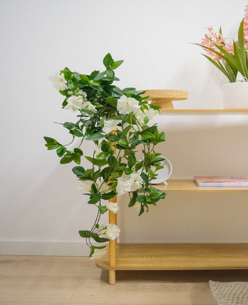 Die künstliche Hängepflanze Petunia 70 cm weiß mit weißen Blüten fällt von einem Holzregal in einen hellen, modernen Raum mit hellen Holzböden. Auf dem obersten Regal steht eine Vase mit langen rosa Blumen.
