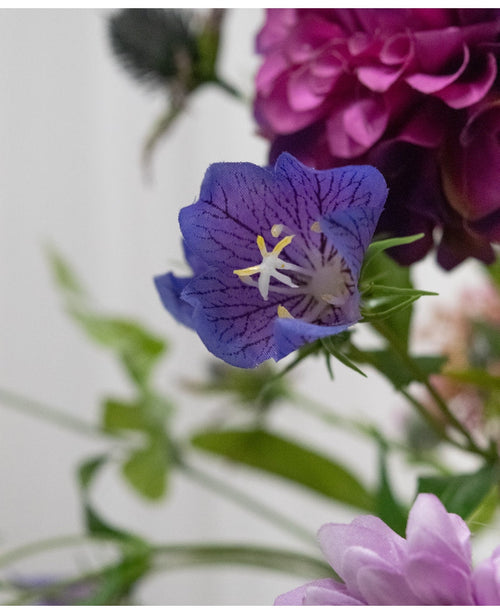 Eine Nahaufnahme der Kunstblume Harebell 62 cm lila, einer zarten lila Kunstblume mit tiefen Adern und gelben Staubgefäßen, zwischen grünen Blättern und zarten zartrosa und violetten Blüten im Hintergrund.