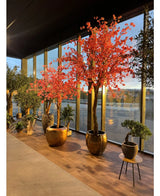 Topfpflanzen und Bäume, wie der 330 cm große künstliche Baum „Pink Cherry Blossom“ mit leuchtend rosa Blüten, sind in der Nähe hoher Glasfenster angeordnet, die das Licht auf dem Holzboden reflektieren.