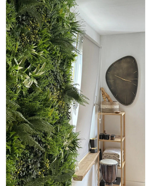 Eine üppige künstliche Hecke Freshness 100x100 cm UV-Vertikalgarten bedeckt eine Wand eines hellen Raumes. Daneben hängt eine moderne dreieckige Wanduhr über einem Holzregal mit Handtüchern, einer Djembe-Trommel, Körben und kleinen Dekostücken.