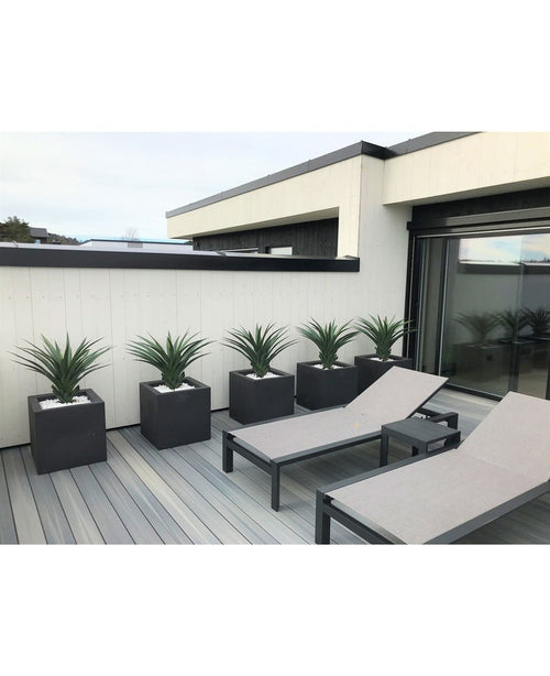 Auf einer modernen Terrasse stehen zwei graue Liegestühle und fünf quadratische Pflanzgefäße, jeweils mit einer künstlichen Pflanze Agave Pandora UV 85 cm, vor einer weiß-schwarz getäfelten Wand für einen eleganten, minimalistischen Look.