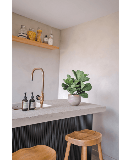 Eine moderne Arbeitsplatte mit einem Messinghahn, zwei Seifenspendern, der künstlichen Tabakpflanze Fiddle Leaf UV 45 cm Topfpflanze und zwei Holzhockern. An einer hellen Wand ist ein schwebendes Regal mit Gläsern und Flaschen montiert.