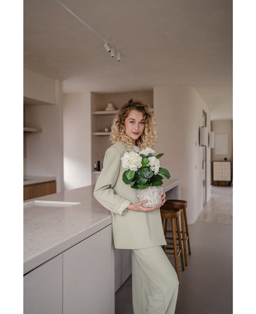 Eine Frau mit lockigen blonden Haaren, gekleidet in ein hellgrünes Outfit, steht in einer modernen Küche mit einer künstlichen Hydrangea White UV 35 cm Pflanze in ihren Händen – mit cremeweißen Blüten – und lächelt in die Kamera.