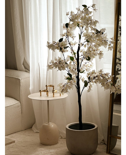 Eine künstliche Bougainvillea-Pflanze mit einer Größe von 120 cm steht neben einem goldumrandeten Spiegel neben einem kleinen runden weißen Beistelltisch mit goldenen Kerzenständern, hohen weißen Kerzen, transparenten weißen Vorhängen und einem cremefarbenen Sofa in einem hellen Raum.