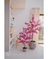 Zwei künstliche Bougainvillea 125 cm Lila mit leuchtend lila Blüten in strukturierten Töpfen erhellen einen modernen Raum neben einem Holzstuhl, einem Bücherregal und einem großen Fenster mit beigen Vorhängen.