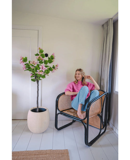 Eine Frau in rosa Pullover und blauen Jeans sitzt barfuß auf einem Rattanstuhl neben der Kunstpflanze Judas 150 cm, einem künstlichen Baum mit rosa Blüten, in einem hellen, modernen Raum mit weißen Wänden und Böden.