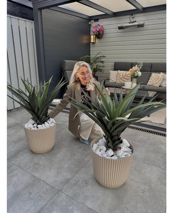 Eine Frau mit langen blonden Haaren kniet und lächelt zwischen zwei künstlichen Agava Pandora UV 85 cm Topfpflanzen auf einer gefliesten Terrasse, mit Gartenmöbeln und Blumen an der Wand im Hintergrund.
