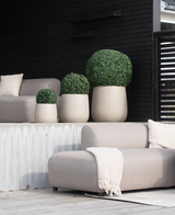 Vier künstliche UV-Buchsbäume im römischen Stil, D40 cm, rund und beige, mit gepflegten grünen Sträuchern, werden auf einer modernen Terrasse mit hellgrauen Stühlen, einer beigen Decke und einem dunklen Holzhintergrund für einen minimalistischen Look platziert.