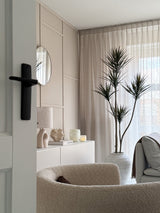 Ein modernes, minimalistisches Wohnzimmer mit einem beigefarbenen Struktursessel, einer künstlichen Pflanze Dracaena Fragnans (180 cm), einem weißen Sideboard mit Dekor, einem runden Spiegel und transparenten Vorhängen, die weiches Tageslicht durch das Fenster filtern.