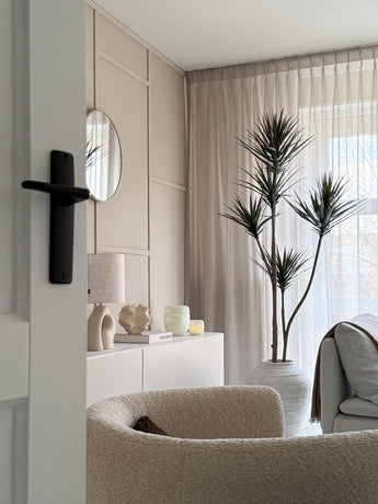 Ein modernes, minimalistisches Wohnzimmer mit einem beigefarbenen Struktursessel, einer künstlichen Pflanze Dracaena Fragnans (180 cm), einem weißen Sideboard mit Dekor, einem runden Spiegel und transparenten Vorhängen, die weiches Tageslicht durch das Fenster filtern.
