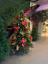 Der künstliche Weihnachtsbaum „Weideman“ PE/PVC – Smart LED steht im Innenbereich in der Nähe einer Treppe, geschmückt mit rosa und roten Blumen, goldenen Blättern und rosa Ornamenten, umgeben von viel Grün auf einem polierten Betonboden für eine stilvolle Weihnachtsdekoration.