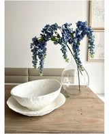 Ein 115 cm großes Blumenarrangement von Kunst Wisteria steht in einer transparenten Vase auf einem Holztisch, daneben zwei weiße Schalen mit Struktur, während die Wand dahinter mit abstrakter Kunst verziert ist.