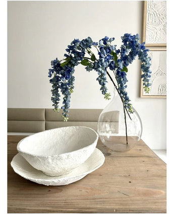 Ein 115 cm großes Blumenarrangement von Kunst Wisteria steht in einer transparenten Vase auf einem Holztisch, daneben zwei weiße Schalen mit Struktur, während die Wand dahinter mit abstrakter Kunst verziert ist.