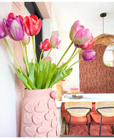 Auf einem Esstisch mit Stühlen mit geflochtenen Rückenlehnen steht eine Vase mit rosafarbenen Herzmotiven mit künstlicher französischer Tulpe 60 cm in Lila. Im Hintergrund tragen ein runder Spiegel und eine braun gemusterte Wand zum gemütlichen, modernen Interieur bei.