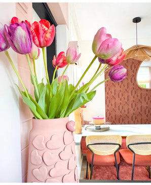 Auf einem Esstisch mit Stühlen mit geflochtenen Rückenlehnen steht eine Vase mit rosafarbenen Herzmotiven mit künstlicher französischer Tulpe 60 cm in Lila. Im Hintergrund tragen ein runder Spiegel und eine braun gemusterte Wand zum gemütlichen, modernen Interieur bei.