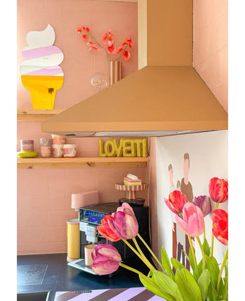 Eine pastellrosa Küche mit künstlicher französischer Tulpe (60 cm) in Lila im Vordergrund, einem gelben LOVETTI-Teller auf dem Regal, rosa Tassen, Wandkunst einer Eistüte und einem minimalistischen Wandgemälde mit zwei Personen an der Wand.