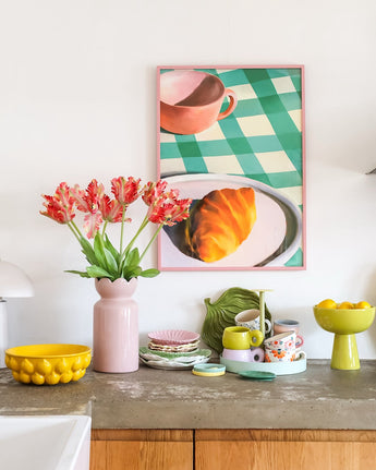 Eine Arbeitsplatte mit allerlei buntem Geschirr und einer künstlichen französischen Sittich-Tulpe von 76 cm in Rot/Gelb in einer rosa Vase. Darüber hängt ein gerahmtes Bild eines Croissants und einer Teetasse auf einer grün karierten Tischdecke.