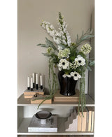 Auf einem Regal an einer hellen Wand steht eine schwarze Vase mit der Kunstblume Amaranthus 124 cm grün, daneben fünf weiße Kerzen, beige Bücher und Dekogegenstände in neutralen Tönen.