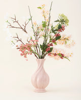 Eine rosa Keramikvase enthält den künstlichen Seidenblumenstrauß Springtime Poem mit weißen, rosa und grünen Blüten mit grünen Blättern und braunen Stielen vor einem hellen Hintergrund.