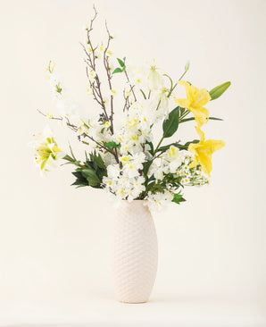 Eine Vase mit weißer Struktur zeigt die Eleganz des künstlichen Seidenblumenstraußes Sunkissed Ivory mit gelben Lilien, weißen Blüten und grünen Blättern vor einem schlichten hellen Hintergrund.