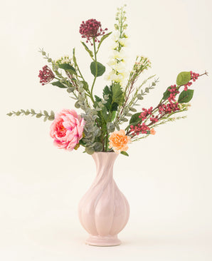 Der Kunstblumenstrauß Pretty Petite aus Seide in einer pastellrosa Vase enthält handgefertigte rosa und orangefarbene Blumen, weiße Blumen, grüne Blätter und rote Beeren – perfekt für eine nachhaltige Inneneinrichtung vor einem schlichten, hellen Hintergrund.