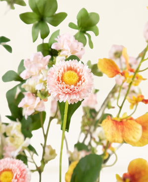 Eine Nahaufnahme des künstlichen Blumenstraußes Blossom Harmony aus Seide mit einer rosa gänseblümchenähnlichen Blume mit einem gelben Herzen, umgeben von hellrosa Blüten und orange-gelben Orchideen, vor einem hellen Hintergrund.