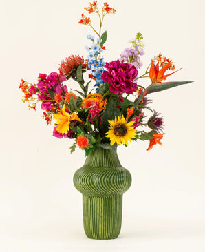 Eine grün strukturierte Vase zeigt den künstlichen Seidenblumenstrauß Colorful Spirit mit Sonnenblumen, rosa Pfingstrosen, orangefarbenen und violetten Blumen und üppigem Grün vor einem schlichten hellen Hintergrund.