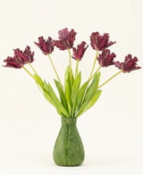 Der künstliche Seidenblumenstrauß „Französische Sittich-Tulpe“ in Lila besteht aus dunkelvioletten Tulpen mit Fransen und langen grünen Stielen in einer Vase mit grünem Blattmotiv, die vor einem weißen Hintergrund eine auffällige Darstellung schaffen.