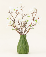 Eine grüne Vase zeigt den Seiden-Kunstblumenstrauß Magnolia weiß mit mehreren weißen Magnolienzweigen mit kleinen grünen Blättern vor einem schlichten hellen Hintergrund.