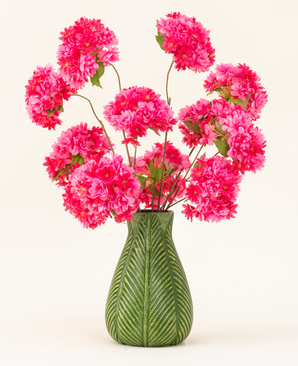 Eine grüne Vase mit Blattmotiv zeigt den künstlichen Seidenblumenstrauß Cherry Blossom Pink – leuchtend rosa, flauschige Seidenkirschblüten mit grünen Blättern – vor einem schlichten hellen Hintergrund.
