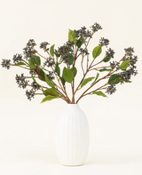 Ein künstlicher Seidenblumenstrauß „Japanese Berry Steel Blue“ mit langen Stielen, dunkelvioletten Beeren und Stahlblättern steht in einer Vase mit weißer Textur vor einem schlichten hellen Hintergrund.