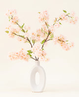 Eine weiße Keramikvase mit einer breiten ovalen Öffnung zeigt mehrere grünblättrige künstliche Seidenblumenstrauß hellrosa Kirschblütenzweige vor einem einfachen hellen Hintergrund.