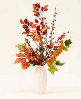 Eine weiße, strukturierte Vase präsentiert den künstlichen Seidenblumenstrauß „Chestnut Charm“ mit Herbstblättern, orangefarbenen Blüten und grünem Blattwerk – ein elegantes künstliches Arrangement vor einem schlichten, hellen Hintergrund.