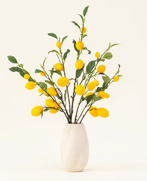 Eine weiße strukturierte Vase zeigt den künstlichen Seidenblumenstrauß „Lemons“ mit grünen Blättern und leuchtend gelben Zitronen vor einem hellen Hintergrund.