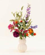 Eine weiße, strukturierte Vase präsentiert den farbenfrohen Kunstblumenstrauß aus Svenski-Seide mit leuchtend lila, orange, rosa und blauen Kunstblumen mit grünen Blättern, die jeden Raum vor einem einfachen hellen Hintergrund erhellen.