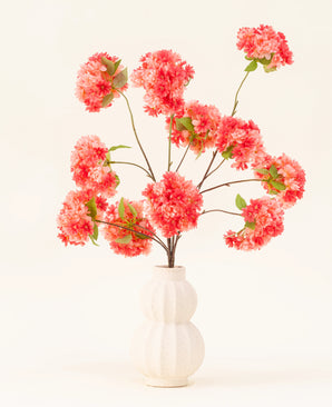 Der künstliche Seidenblumenstrauß Cherry Blossom Coral besteht aus leuchtend rosa und korallenroten künstlichen Kirschblüten mit grünen Blättern, elegant arrangiert in einer weißen, geformten Vase vor einem hellen Hintergrund.