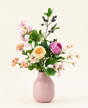 Der künstliche Blumenstrauß Touch of Cloudlight aus Seide, präsentiert in einer rosa Keramikvase, enthält rosa Pfingstrosen, gelbe Rosen, kleine Blüten, Grünpflanzen und Zweige mit dunklen Beeren vor einem hellen Hintergrund.