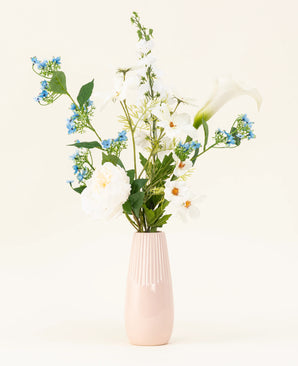 Die hellrosa Vase enthält den Kunstblumenstrauß Whispers of White aus Seide mit weißen Blumen, blauen Blüten und grünem Blattwerk vor einem schlichten, hellen Hintergrund.