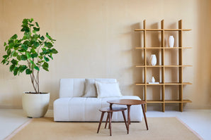 Minimalistisches Wohnzimmer mit einem weißen Sofa, zwei ineinander gesteckten Couchtischen aus Holz, beigen Wänden, einem offenen Holzregal mit weißen Vasen und einer Kunstpflanze Ficus 180 cm, die für viel Grün sorgt. Ein beiger Teppich bedeckt den Boden.