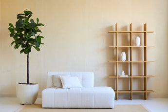 Minimalistisches Wohnzimmer mit einem weißen Sofa, zwei Kissen und einer Kunstpflanze Ficus Tabakpflanze Deluxe 180 cm neben einem offenen Holzregal mit weißen Vasen, alles vor einer beigen Wand und auf einem hellen Teppich.