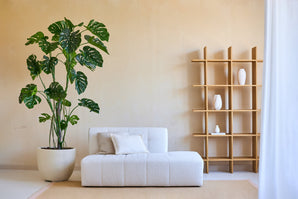 Minimalistisches Wohnzimmer mit einer künstlichen Monstera-Pflanze 240 cm in einem Topf, einem modernen weißen Sofa mit Kissen und einem offenen Holzschrank mit weißen Vasen vor einer beigen Wand. Natürliches Licht von rechts.